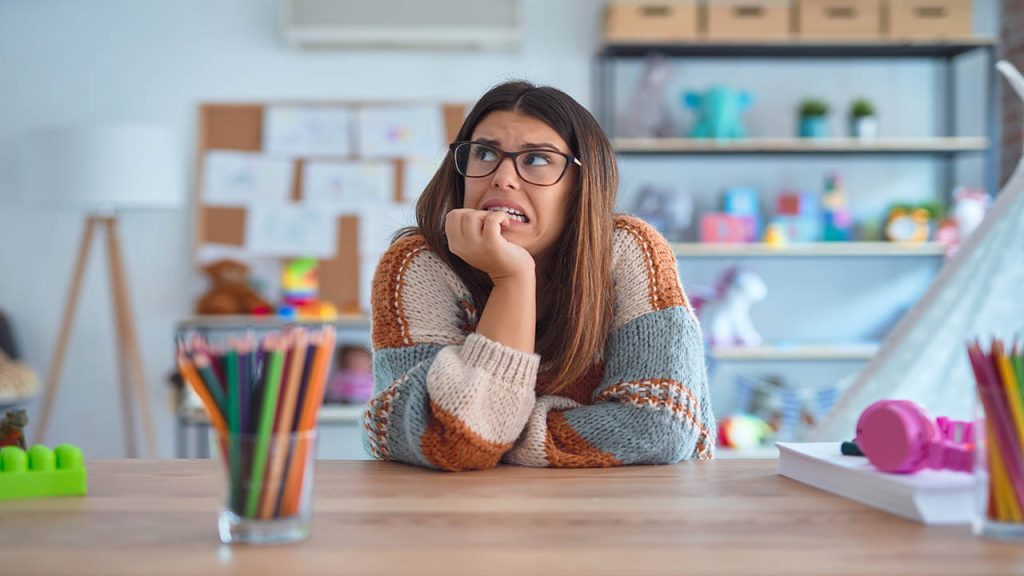 Young beautiful teacher woman wearing sweater and glasses sitting on desk at kindergarten looking stressed and nervous with hands on mouth biting nails. Anxiety problem.