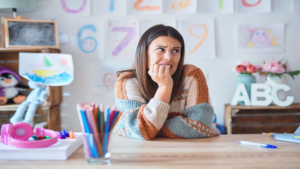 Young beautiful teacher woman wearing sweater and glasses sitting on desk at kindergarten looking stressed and nervous with hands on mouth biting nails. Anxiety problem.