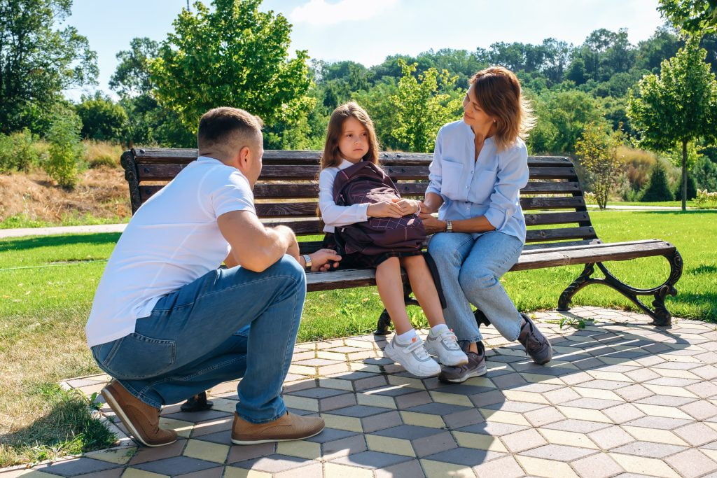 Unhappy schoolgirl in uniform and with backpack and sits with parents on a bench in the park and does not want go to school