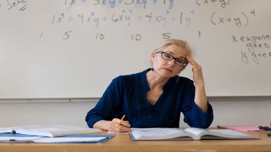 Portrait of frustrated tired high school math teacher sitting on desk talking to students.