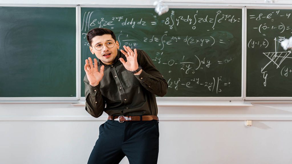 crumpled paper balls flying at frightened male teacher in classroom with chalkboard on background