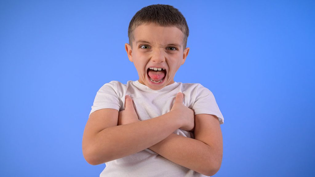 Portrait of aggressive boy with arms crossed shouting and standing on isolated blue background