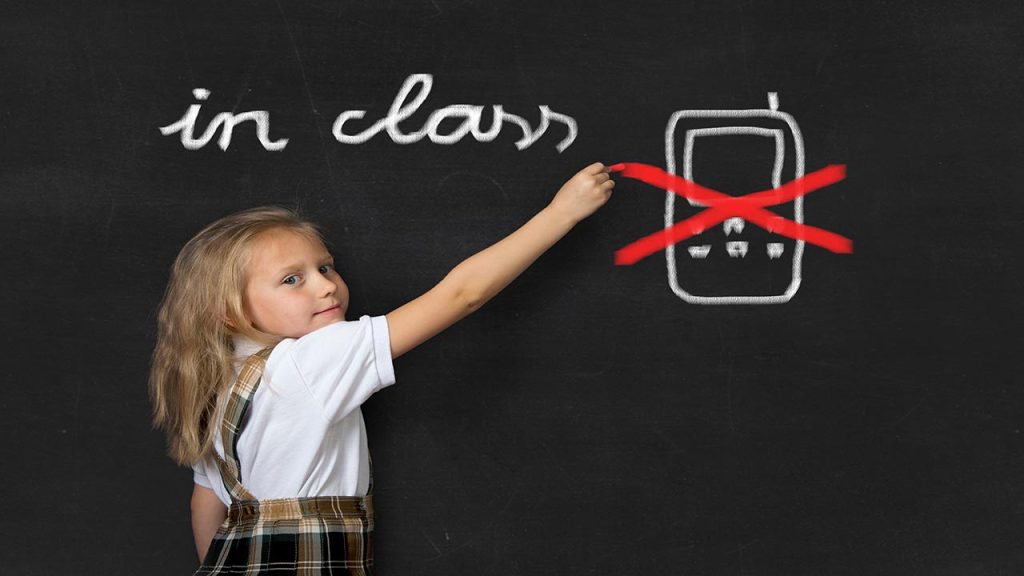 young sweet junior schoolgirl with blonde hair standing happy and smiling writing with chalk about not using mobile phone in school class in education rules and cellular forbidden concept